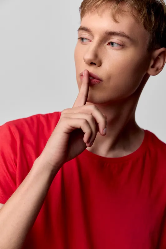 Young man in a red shirt signals silence with a finger to lips, conveying secrecy, hush, and quiet focus. A minimalist studio shot emphasizes mood, contemplation, and restrained communication against
