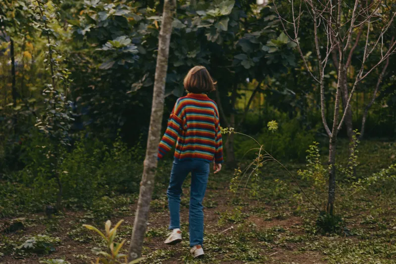 Child kid walking in forest nature wearing colorful striped sweater, back view of kid exploring a woodland path in jeans and sneakers, candid outdoor scene with soft autumn light.
