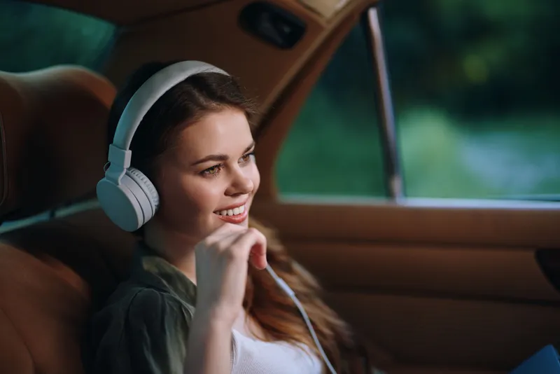 Young woman enjoying music in a car, wearing headphones, smiling, with a natural outdoor background, embracing a carefree and relaxed summer vibe.