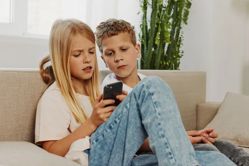 Two focused children sitting on sofa using smartphone together, sharing digital content. Cozy home interior with natural light and green plant. Concept of technology, childhood, and learning.
