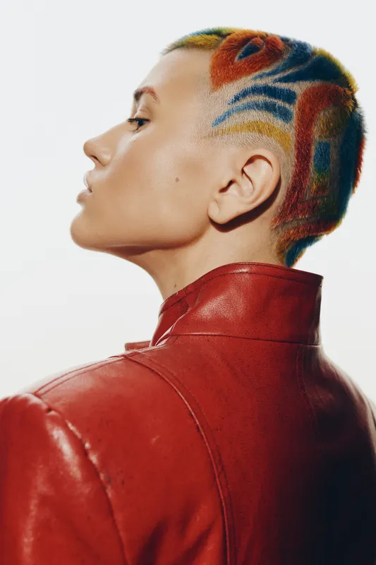 Woman with colorful patterned shaved hair wearing red leather jacket, showcasing bold style and artistic hair design in a close-up profile portrait.