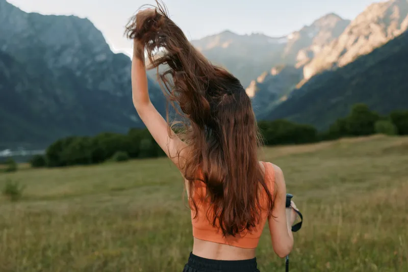 A woman with long hair in an orange tank top stands in a grassy meadow with distant mountains, lifting her hair while holding a camera, enjoying a bright outdoor scene.