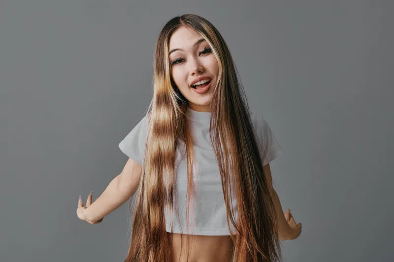 Teen girl with long burgundy ombre hair smiling and posing with arms outstretched on gray background in studio. Young woman with stylish hairstyle and casual white t shirt.