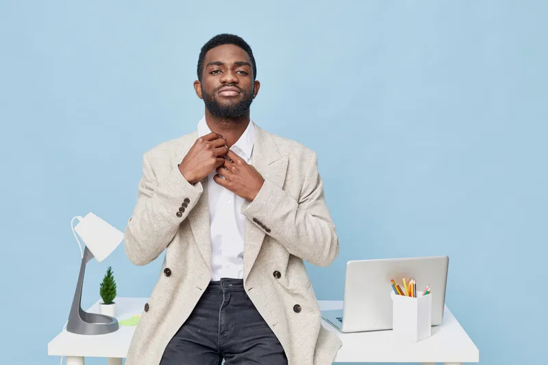 Confident young African American man in beige blazer and white shirt adjusting collar while standing at modern workstation with desk lamp, laptop, and stationery on light blue background. Business sty