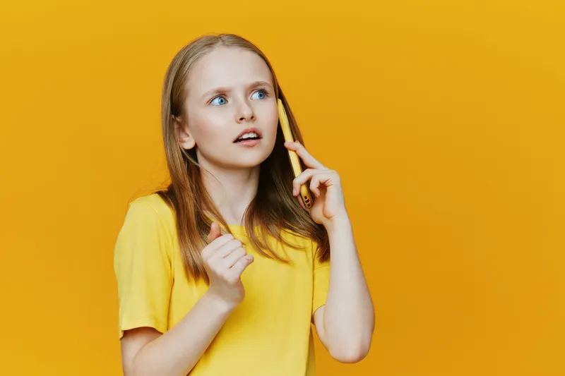 Surprised girl talking on the phone against a vibrant yellow background, showcasing her youthful style and expression of curiosity