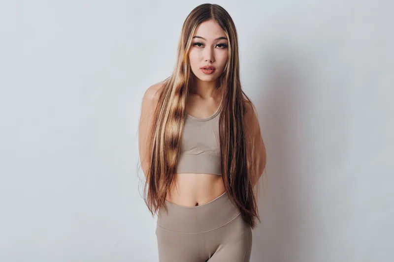 Young woman with long straight hair wearing fitness clothes posing indoors with neutral expression on white background. Female model in beige activewear standing front view with arms behind back.