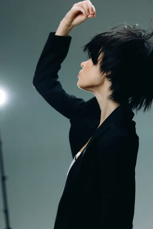 Stylish woman in black jacket posing with raised arm against gray background in studio lighting