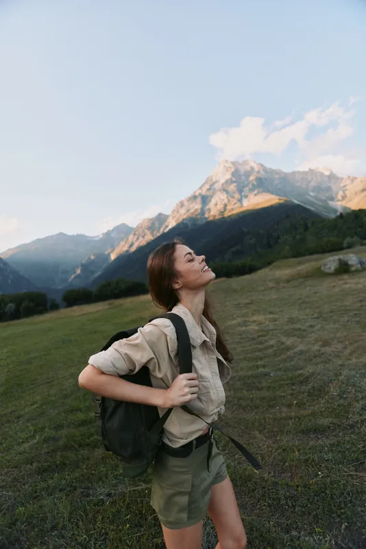 Young woman hiking through a mountain meadow with a backpack, looking up at the peaks with a bright carefree smile under clear blue skies.