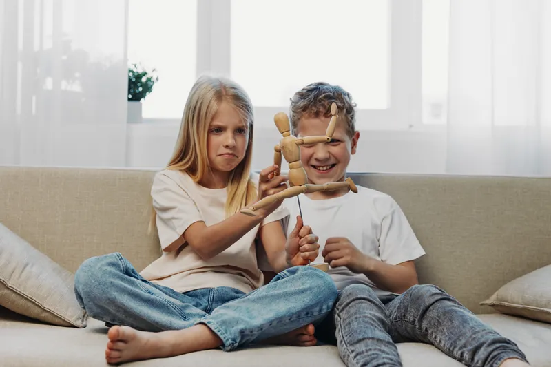 Two children sitting on couch play with wooden mannequin, girl looks curious and boy smiles in bright living room with casual clothes and natural light.