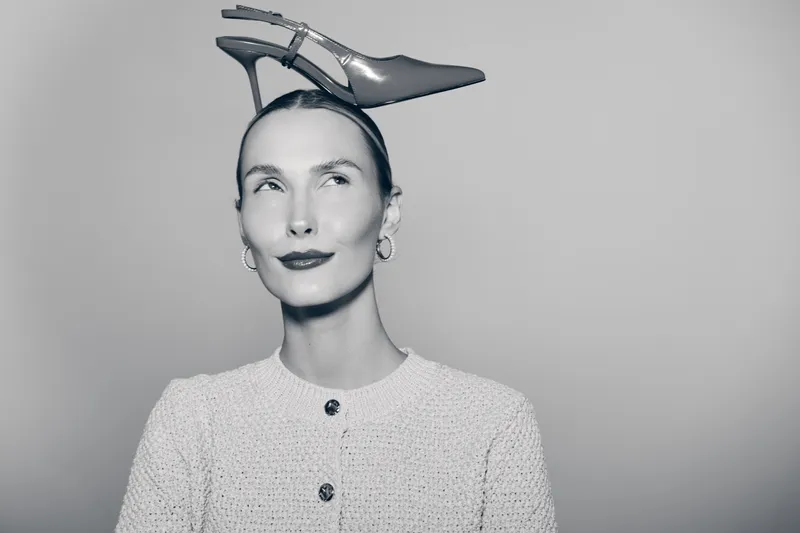 Portrait of a fair skinned Caucasian woman with slicked hair balancing a shoe on her head, wearing earrings and a textured buttoned top in black and white.