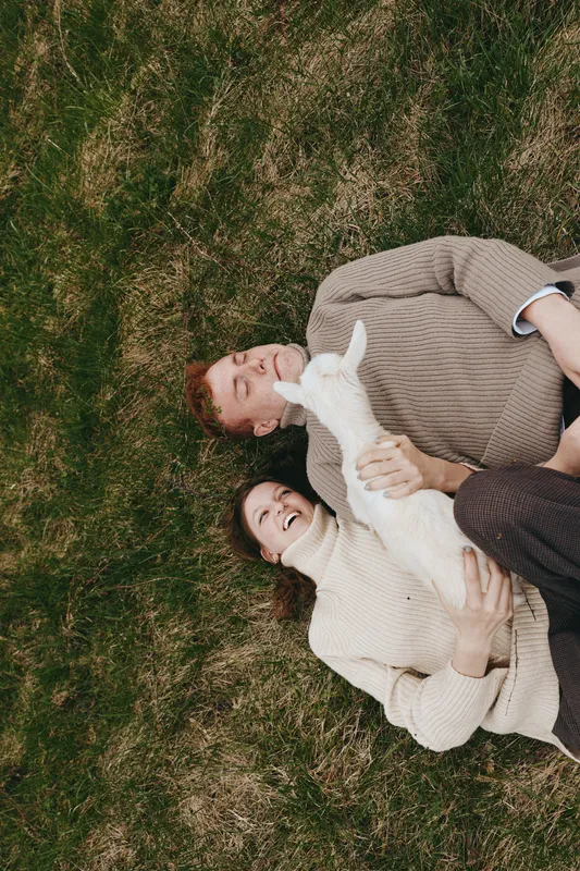 Cheerful couple outdoors, lying on grass with a goat kid nearby, warm sunlight, relaxed posture, candid moment, rustic setting, natural tones, gentle breeze, wholesome lifestyle scene