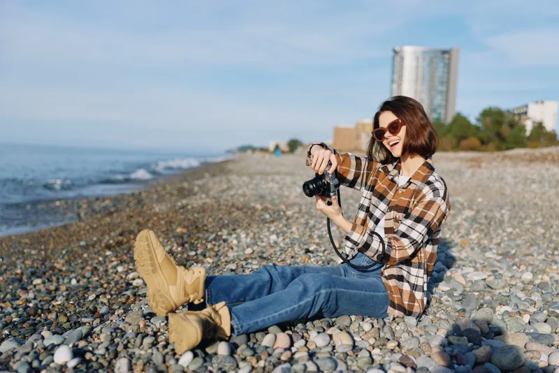Photographer sitting on a pebble beach with a camera, wearing a plaid shirt, jeans and boots, enjoying a sunny day by the sea with distant buildings and a blue sky.