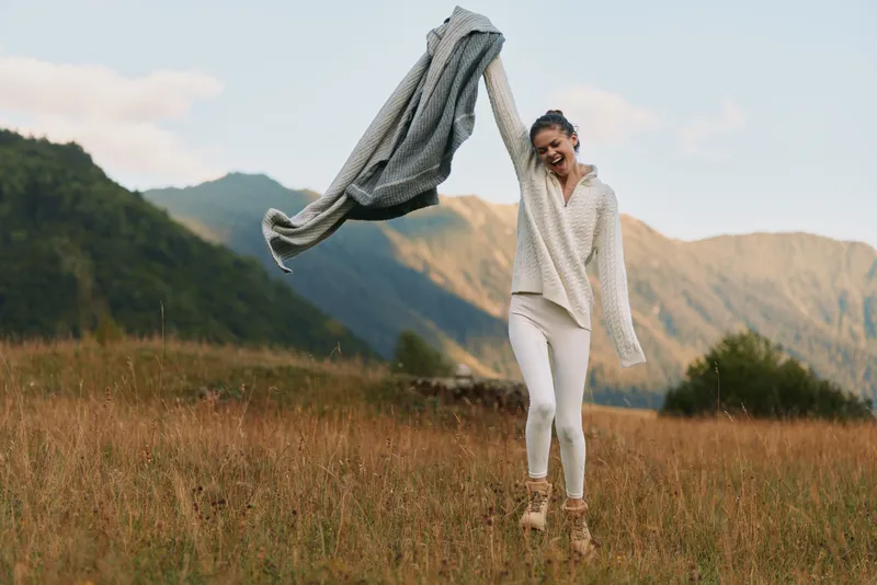 A woman enjoys outdoor lifestyle in a grassy field with mountains in the background, raising a scarf as she smiles, conveying freedom and carefree weekend mood