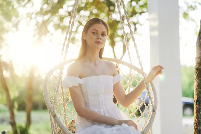 Young woman in a white dress sitting on a swing in a sunlit garden, showcasing serenity and elegance Ideal for themes of nature, beauty, and tranquility