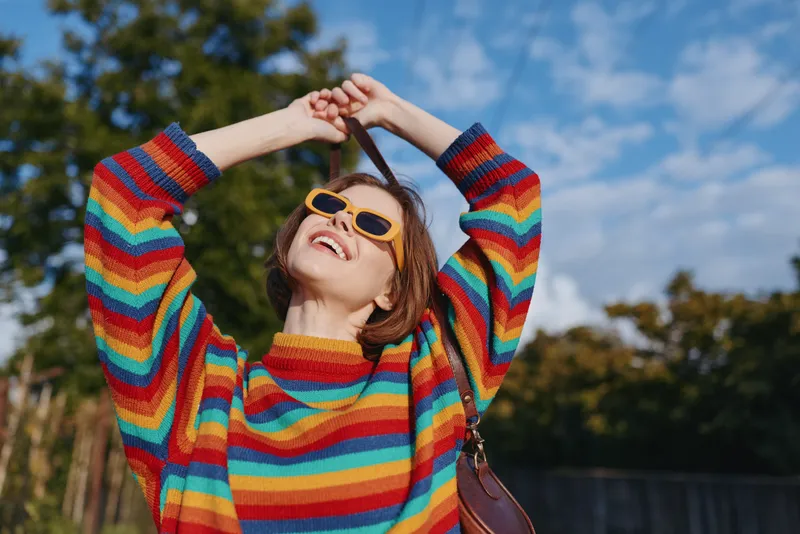 Woman in a rainbow sweater wearing yellow sunglasses, smiling and joyful outdoors in a park. Young adult with casual outfit and shoulder bag posing carefree under blue sky.