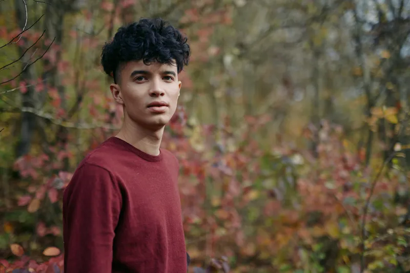 Close-up portrait of a young man standing among autumn foliage, natural light capturing a genuine expression, textured sweater and soft bokeh that emphasize authenticity and steady credibility.