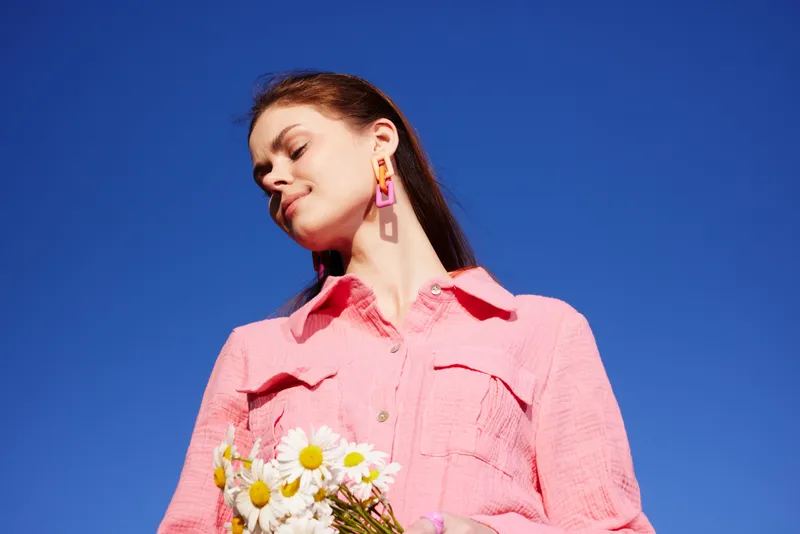 Young woman smiling joyfully, wearing a pink blouse, holding daisies against a clear blue sky, capturing a vibrant spring mood, ideal for lifestyle and wellness themes