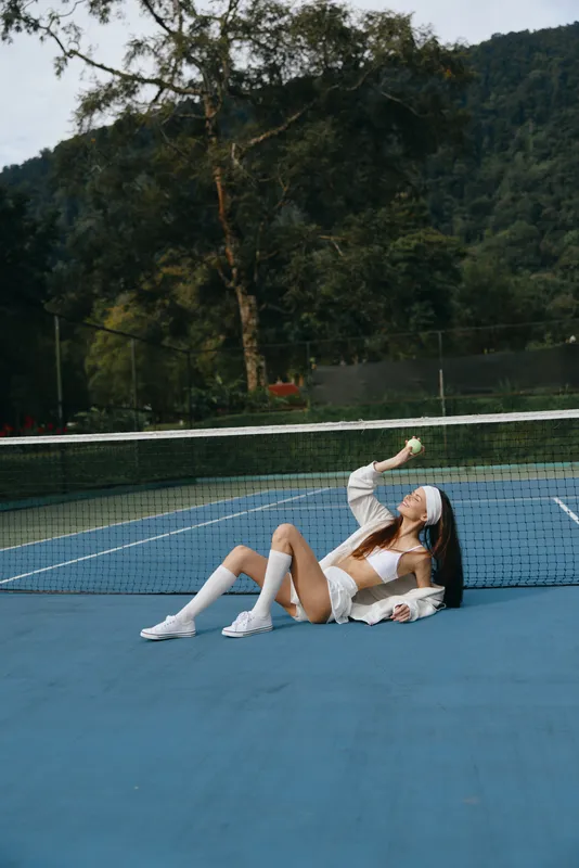 Young female tennis player in sportswear expressing joy on the court, surrounded by lush greenery and a vibrant blue surface, perfect for healthy lifestyle and sports photography