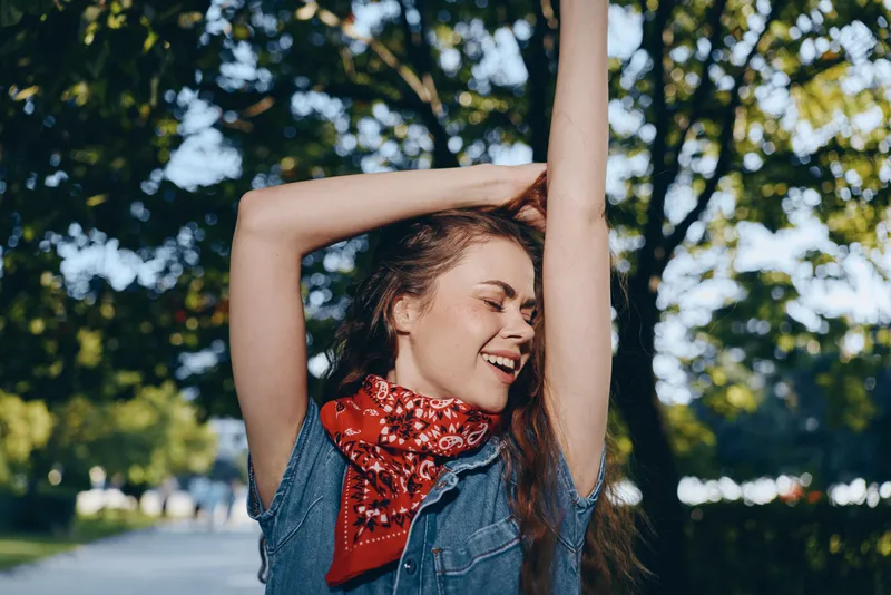 Smiling woman outdoors with raised arms wearing casual denim and red scarf enjoying sunny day in green park surrounded by trees with joyful expression.