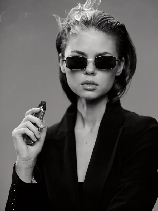 Black and white portrait of a stylish woman in sunglasses holding a vape, exhaling smoke, wearing a black blazer with a confident expression.