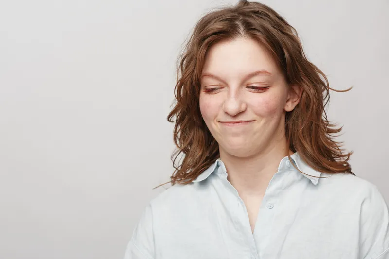 Young woman with wavy hair and white shirt makes a funny face with closed eyes and slight smile on light background. Concept of humor, emotion, and casual portrait.