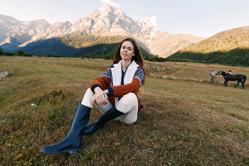 Woman sits in mountain meadow wearing sweater and boots, enjoying countryside nature with horses grazing in background, autumn light and relaxed outdoor lifestyle portrait.
