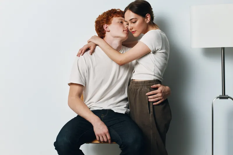 Couple in intimate embrace with woman standing and man sitting on stool against white wall with floor lamp, showing affection and closeness in cozy indoor setting.