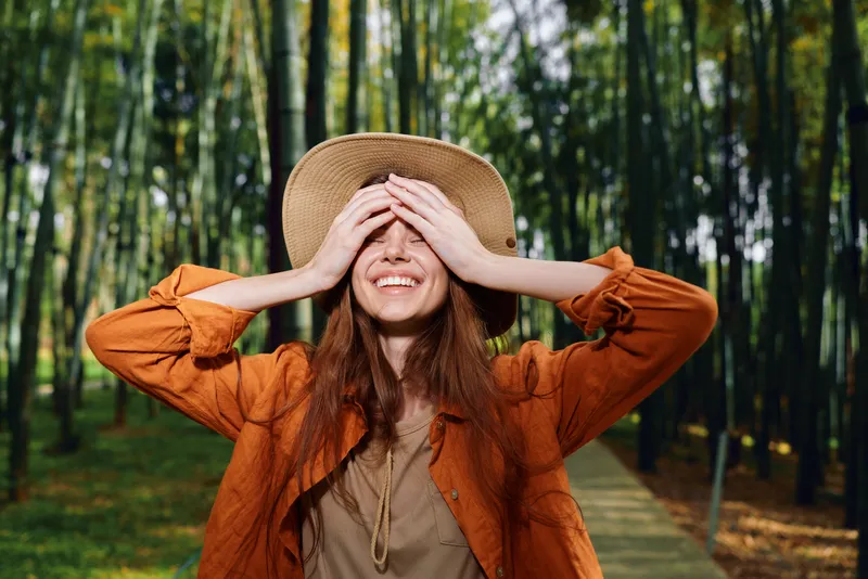 Woman in straw hat smiling with her hands covering her eyes on a bamboo forest path, wearing an orange jacket and enjoying a happy, carefree outdoor portrait in nature.