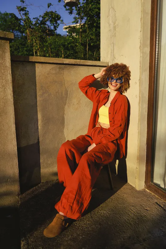 Young woman sits on a sunny balcony wearing a red tracksuit and yellow top, shielding eyes with sunglasses and smiling, enjoying warm daylight