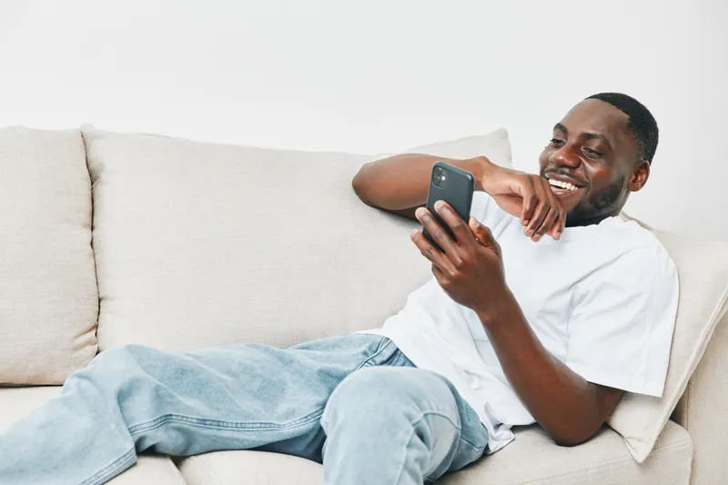 Smiling African man relaxing at home, browsing on smartphone while sitting on couch, wearing casual white t shirt and blue jeans, demonstrating happiness and comfort