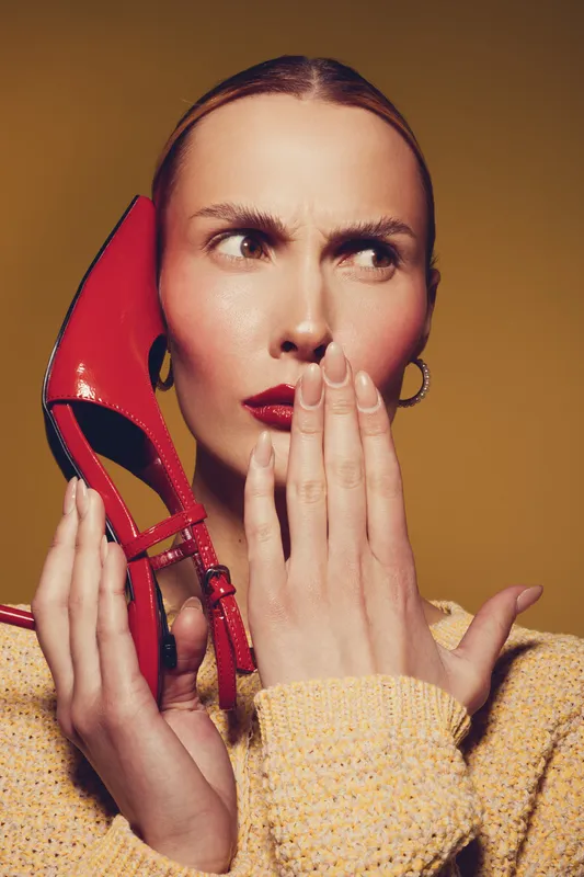 Curious woman in yellow sweater holds red high heel shoe near face, covering mouth with hand. Concept for fashion, surprise, and style expression in studio lighting.