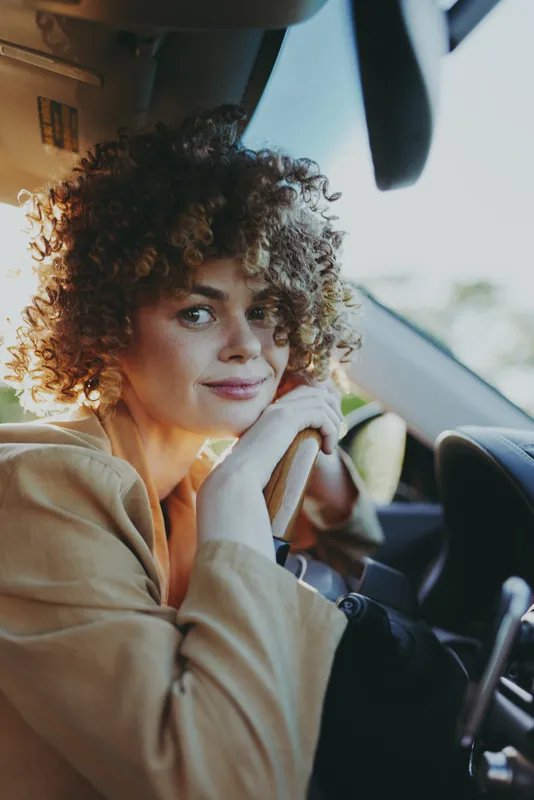 Smiling woman with curly hair sits in a car, looking at the camera. Warm light fills the cabin, creating a cozy, relaxed mood and casual style. Theme of daily travel and self care.