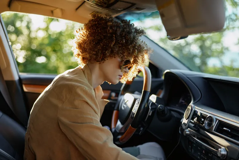 Curly haired woman driving a car, focused on the road, warm retro film color palette, cozy interior, everyday lifestyle mood, candid moment, soft lighting and thoughtful expression.