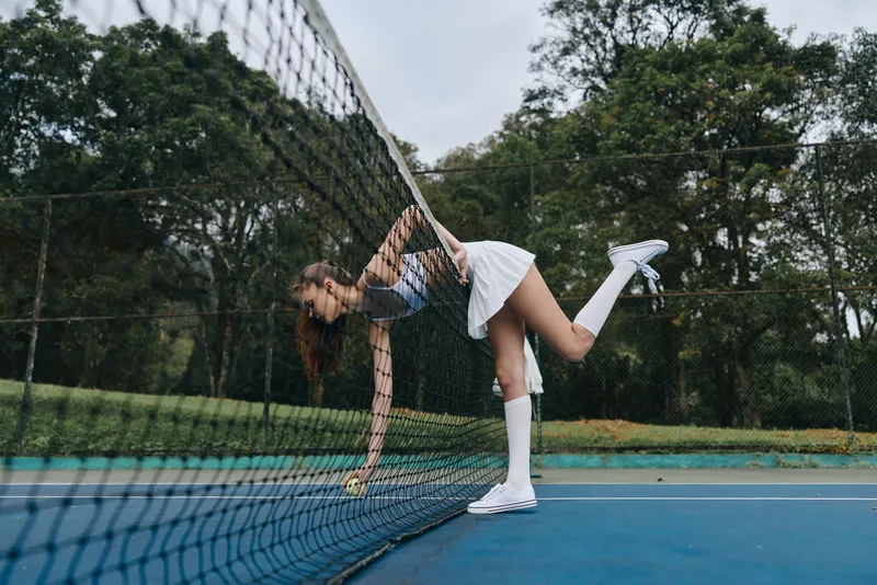 Young athletic woman in a tennis outfit reaching for a ball on a court, showcasing determination and sportiness amidst a green backdrop