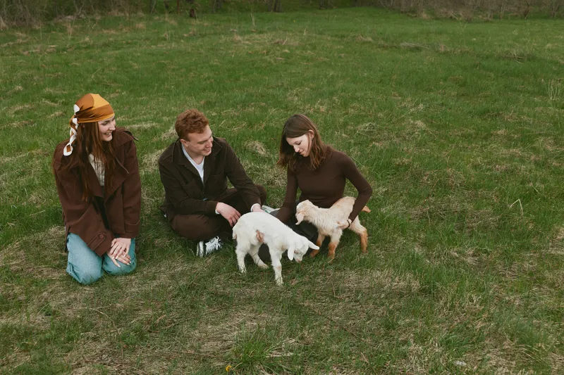 Three friends interact with a shy goat and its kids on a grassy field, sharing smiles and gentle petting, capturing a candid lifestyle moment in a rural setting, warm and sunny