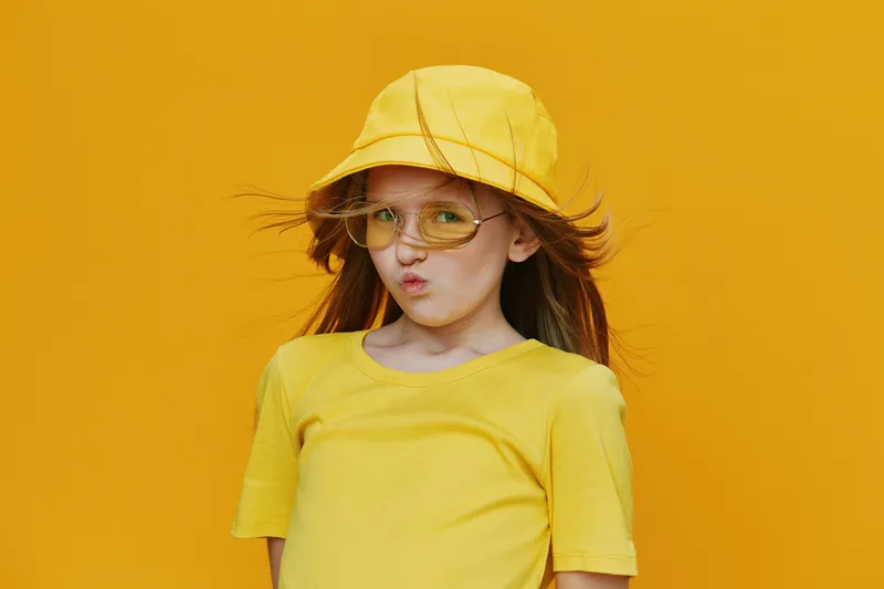 A cheerful young girl in a yellow outfit with a matching bucket hat and sunglasses poses playfully against a vibrant yellow background, radiating summer vibes and joy