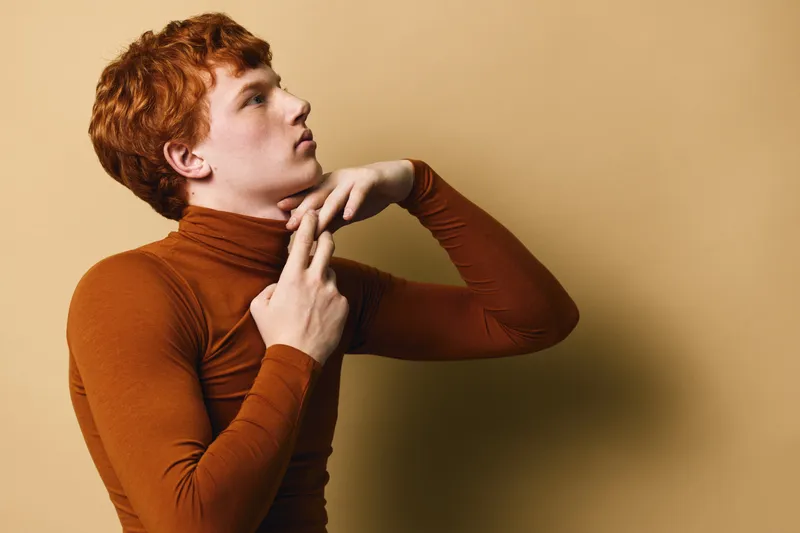 Redhead man wearing brown turtleneck posing and looking sideways on beige background with thoughtful expression and hand near neck in studio portrait.