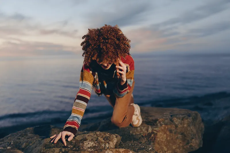 Woman lifestyle outdoors by the sea, crouching on a rock at sunset, curly hair framing her face, one hand touching hair, calm ocean waves behind, cinematic mood, soft light, evening clouds