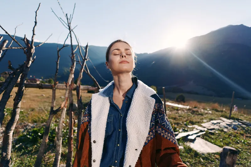 Woman standing in mountains with sunlit outdoors scene, enjoying nature in a meadow wearing a warm jacket, eyes closed in relaxation and peaceful outdoor portrait at golden hour