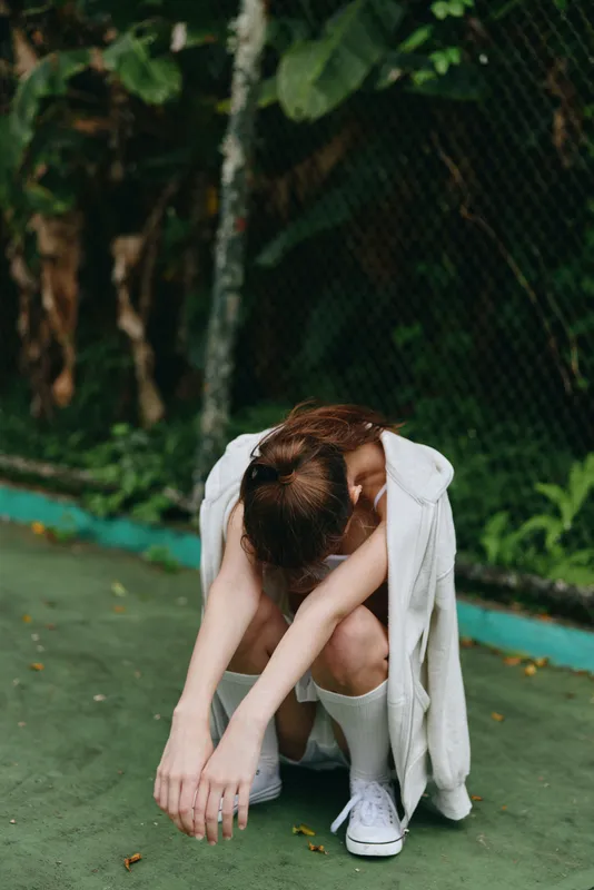 A young woman with long brown hair, dressed in a white hoodie and knee high socks, reflects in a moment of contemplation on a green urban court surrounded by lush foliage