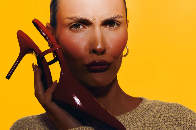 Close-up of serious woman holding red high heel shoe near face on bright yellow background. Concept of fashion, style, footwear, and bold color contrast in studio lighting.