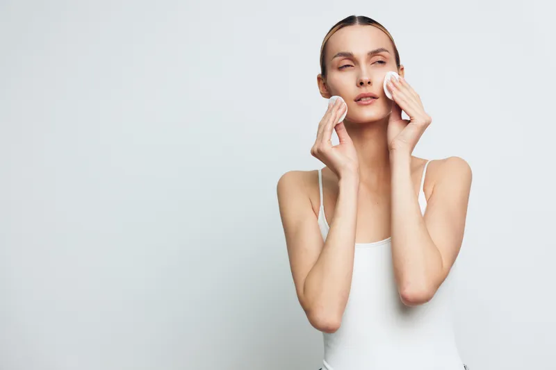 Young woman cleanses her face with cotton pads in a bright studio, showing calm focus and a refreshing skincare moment with soft lighting and a clean backdrop