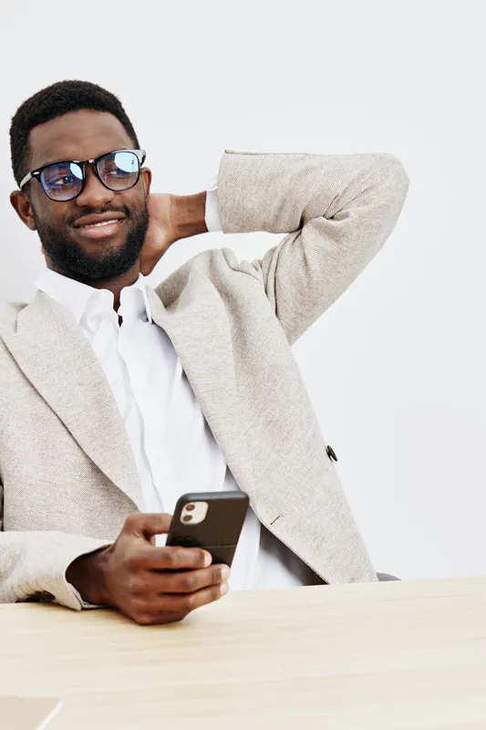 Smiling man wearing glasses and beige blazer relaxed while using smartphone sitting at desk isolated on plain white background studio portrait. People lifestyle concept