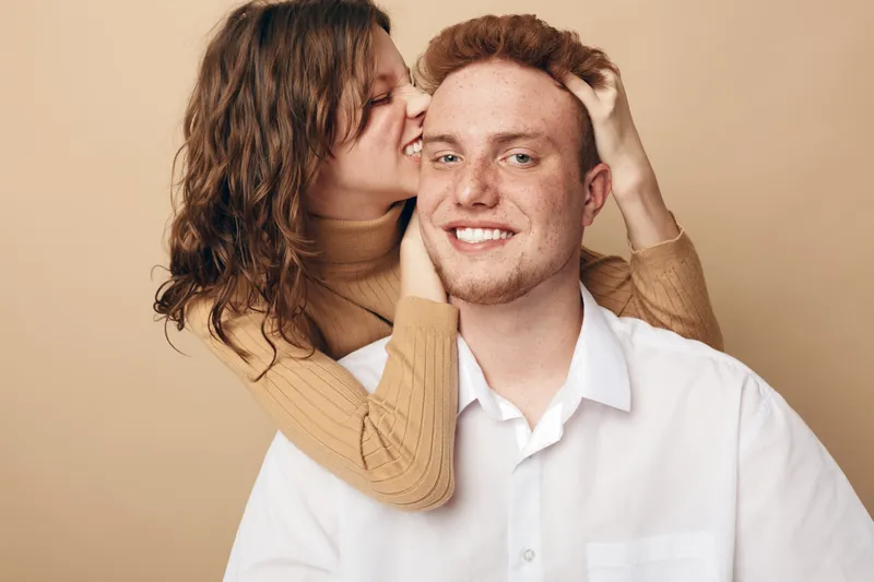 Happy young couple in casual clothes share intimate moment, woman whispering in man’s ear with smile. Beige background, warm lighting, concept of love, affection, and close relationship.
