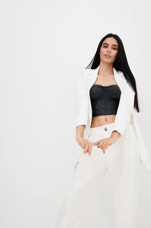 young woman wearing stylish black bustier and loose white pants with matching blazer posing confidently against plain white background in modern fashion look