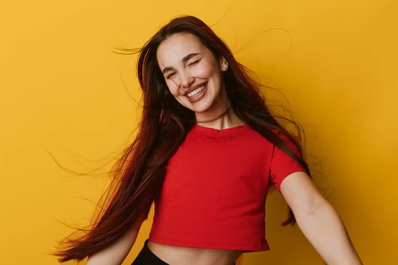Young woman with long hair smiling and winking in red crop top against yellow background. Happy female portrait with natural makeup and casual style for positive emotion and youth.