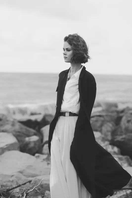 Woman on a rocky coast wearing a long coat and flowing dress, wind swept hair, moody grayscale scene, minimalist styling, reflective mood, seaside fashion, serene yet powerful