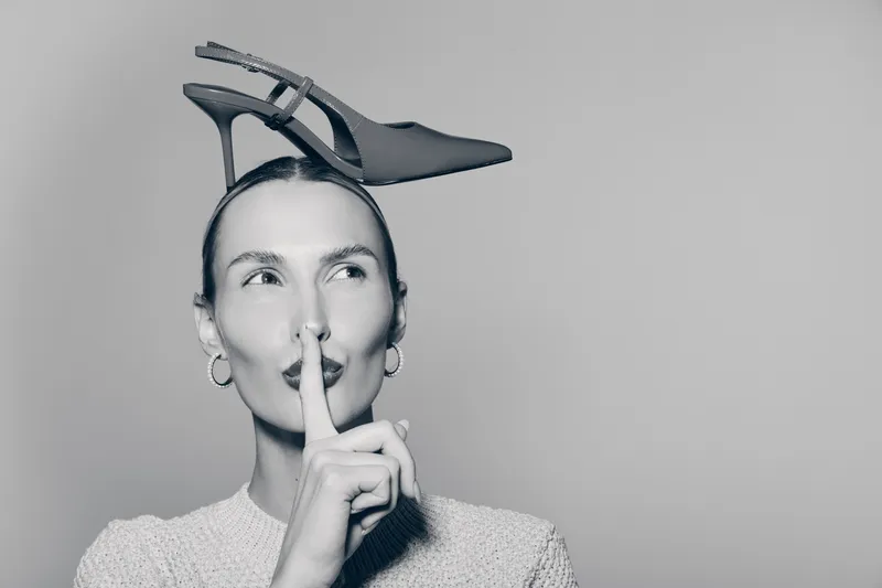 Young white woman with fair skin holding finger to lips and black high heel shoe on head. Concept for silence, fashion, and playful mood.