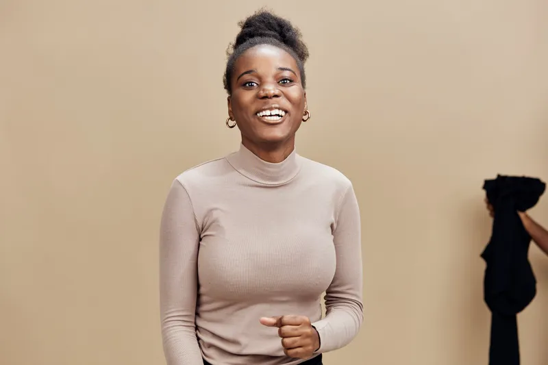Happy young African woman wearing a beige turtleneck smiles against a neutral background, embodying confidence and joy in a modern lifestyle concept