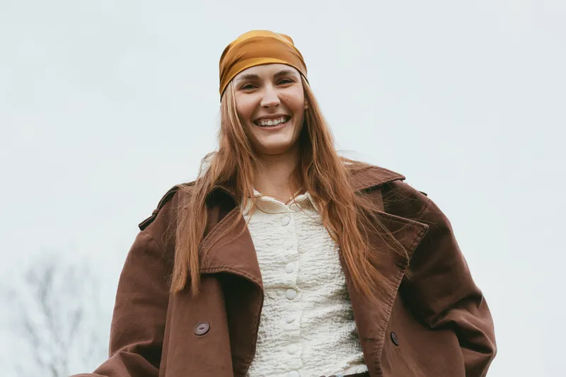 Cheerful woman outdoors, smiling, wearing a warm brown coat and bandana, casual pose, natural colors, soft breeze, sunny atmosphere, candid lifestyle portrait, autumn tones, relaxed vibe, simple